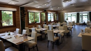 A spacious restaurant with multiple tables and chairs, large windows with greenery visible outside, and a clean, modern interior design. - Logis Hôtel Arc en Ciel (Thonon-les-Bains)