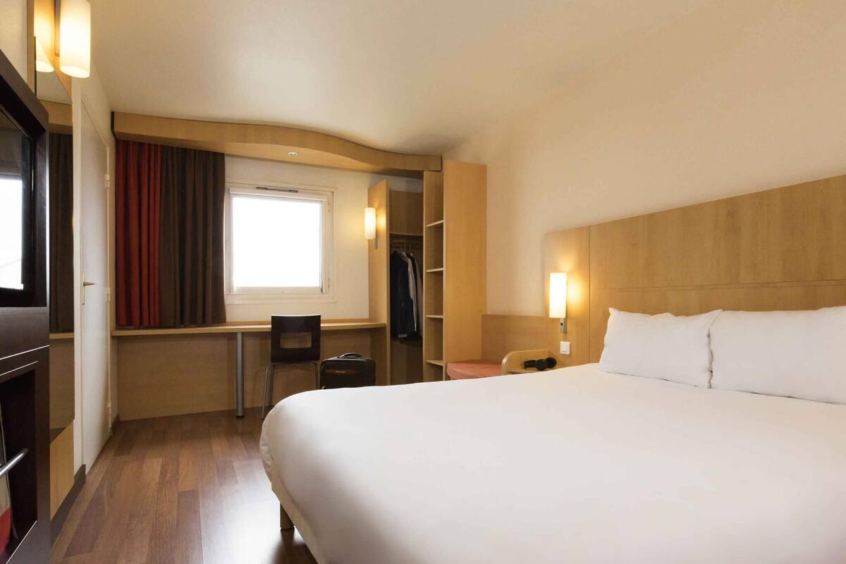 Habitación de hotel con una cama grande, un escritorio con silla, un armario y una ventana con cortinas.