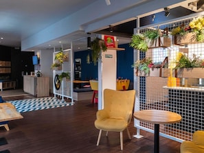 A modern interior with a wooden reception desk, a yellow chair, a round table, and a blue wall with a geometric pattern. - ibis Lille Lomme Centre (Lille)