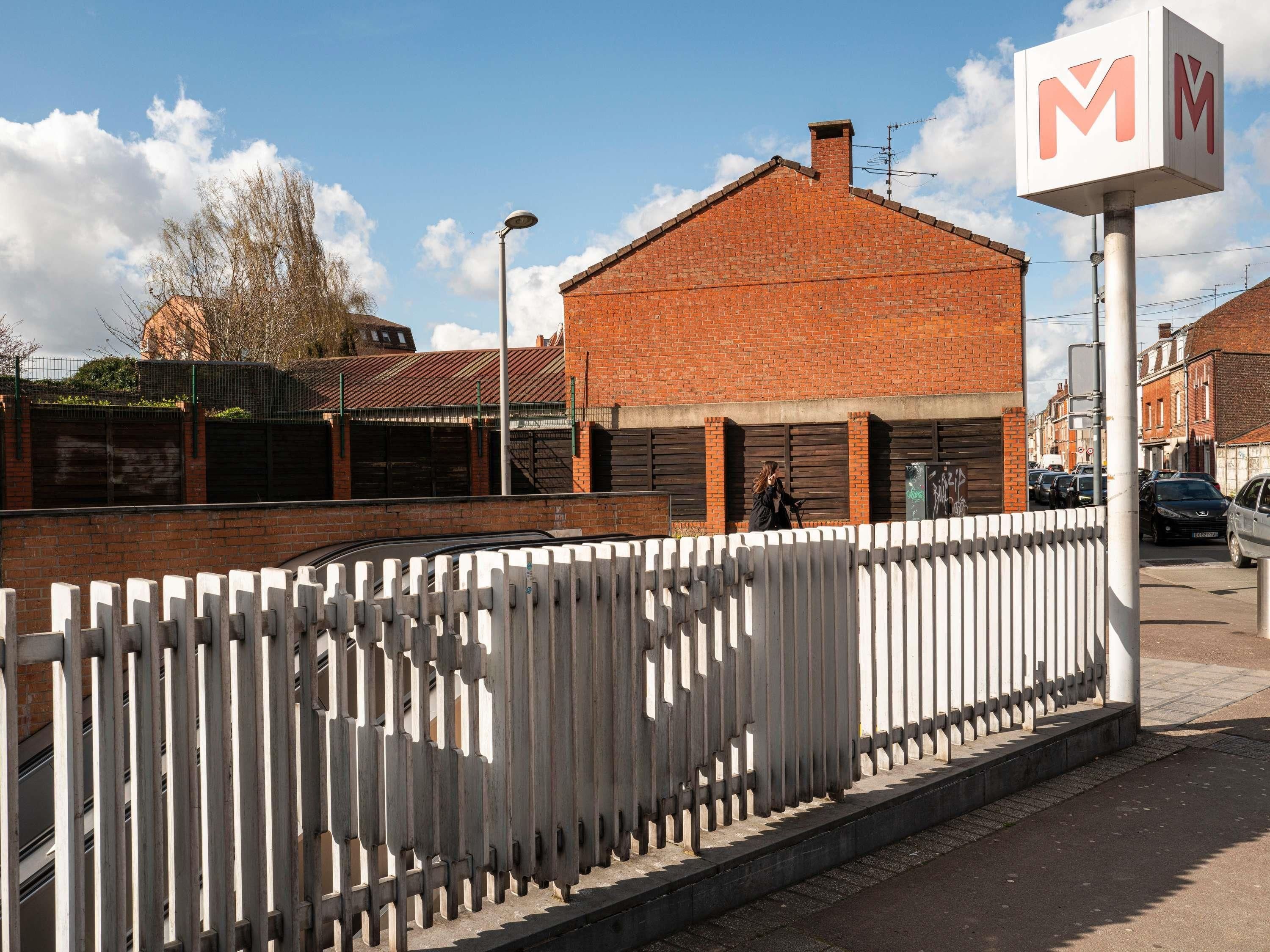 A brick building with a white fence, a street lamp, and a sign with the letter 'M'.