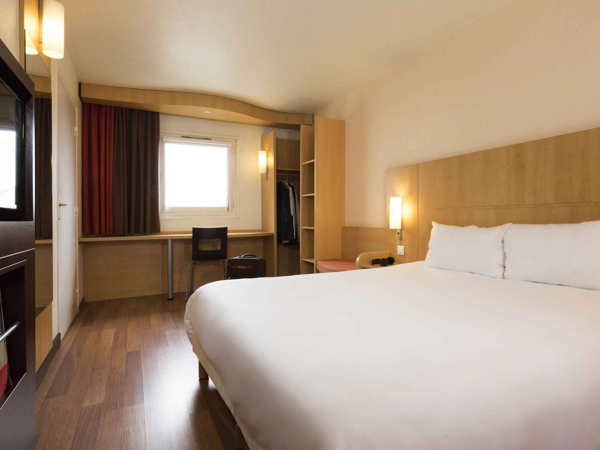 A hotel room with a large bed, a desk with a chair, a wardrobe, and a window with curtains.