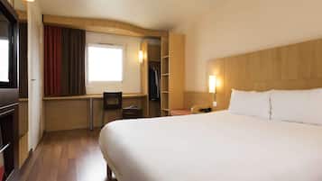 A hotel room with a large bed, a desk with a chair, a wardrobe, and a window with curtains.