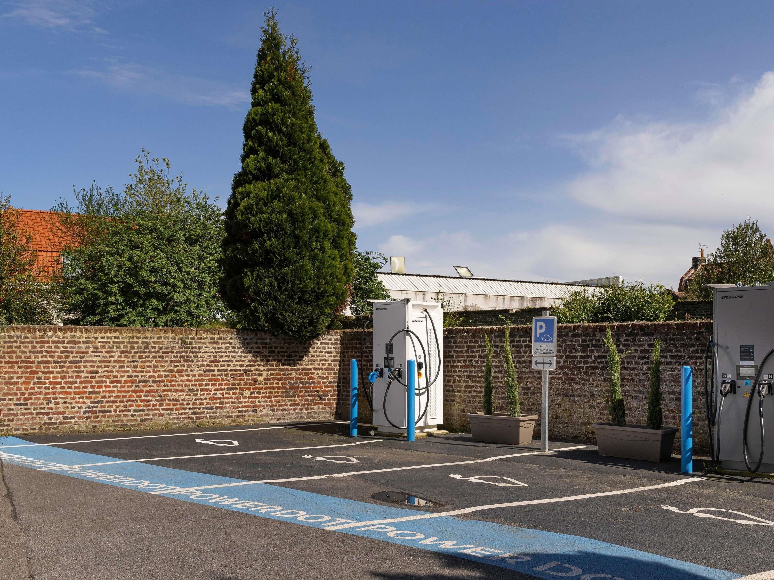 Un área de estacionamiento con estaciones de carga para vehículos eléctricos, un muro de ladrillo y un gran árbol.