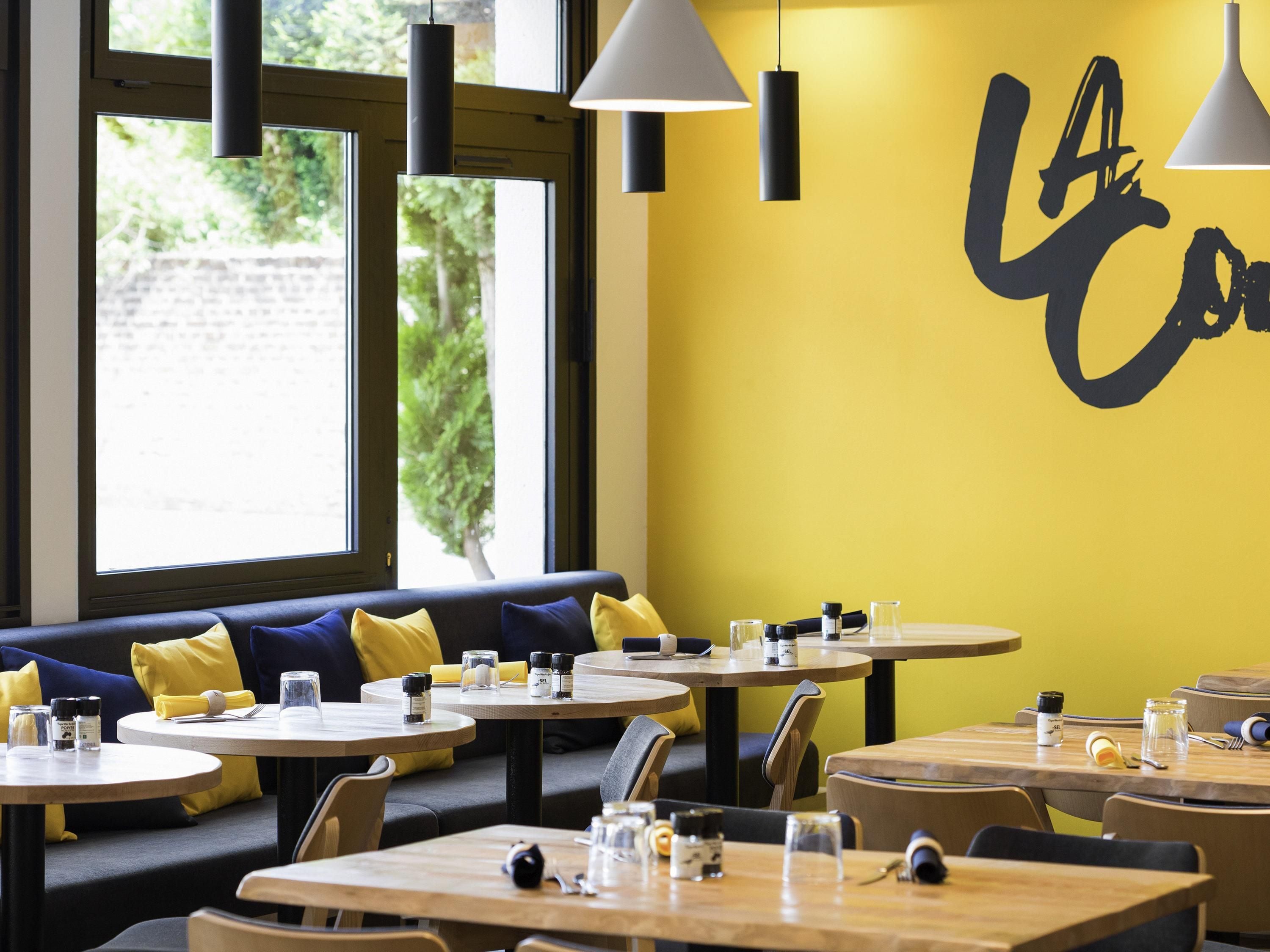 A restaurant interior with round tables, yellow cushions, and a wall with a large black letter design.