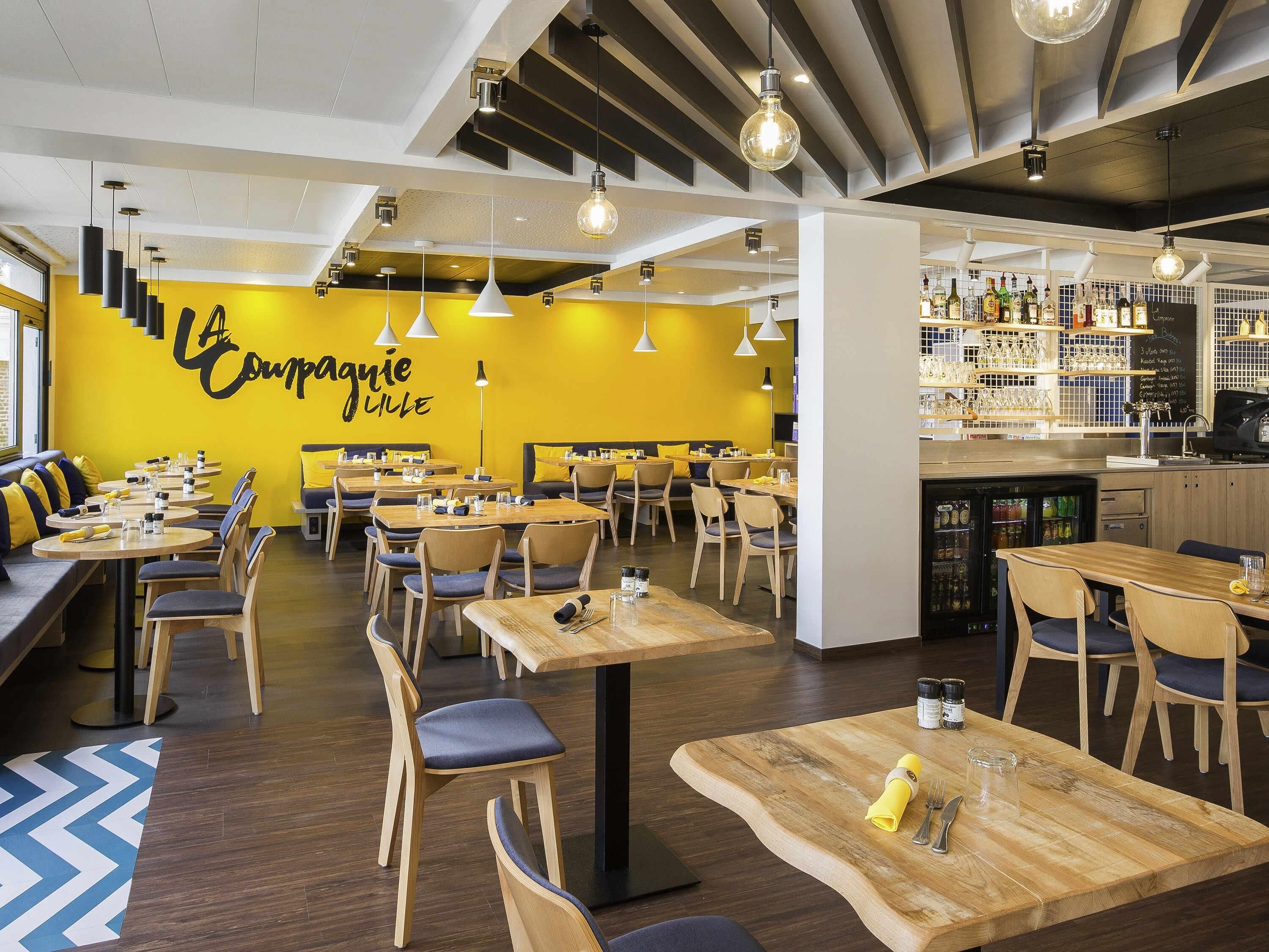 A restaurant interior with multiple dining tables and chairs, a bar area with shelves of bottles, and a yellow wall with a sign.