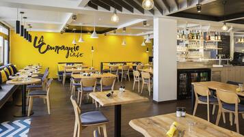 A restaurant interior with multiple dining tables and chairs, a bar area with shelves of bottles, and a yellow wall with a sign.