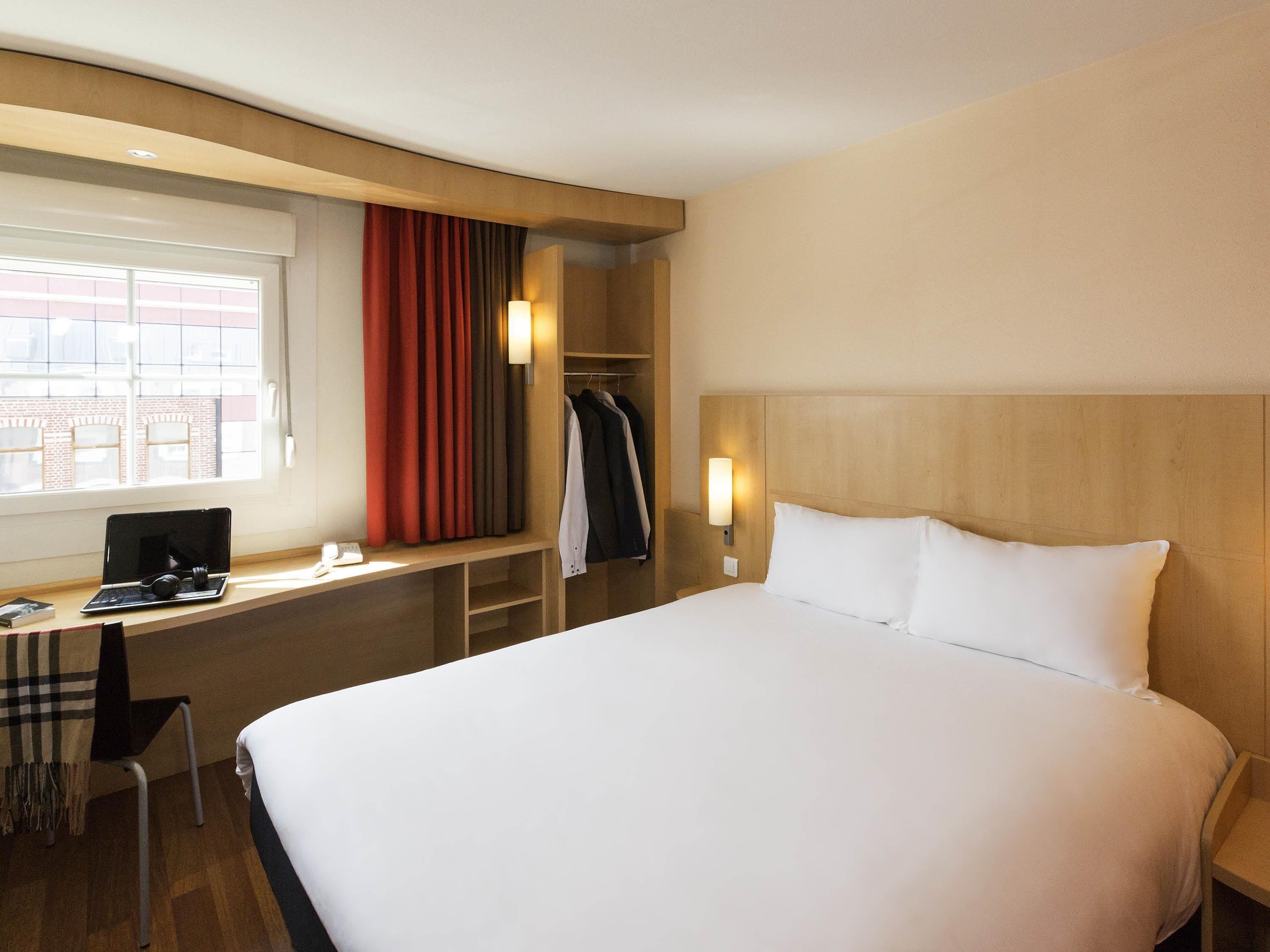 A hotel room with a bed, a desk with a laptop, a chair, a coat rack, and a window with curtains.