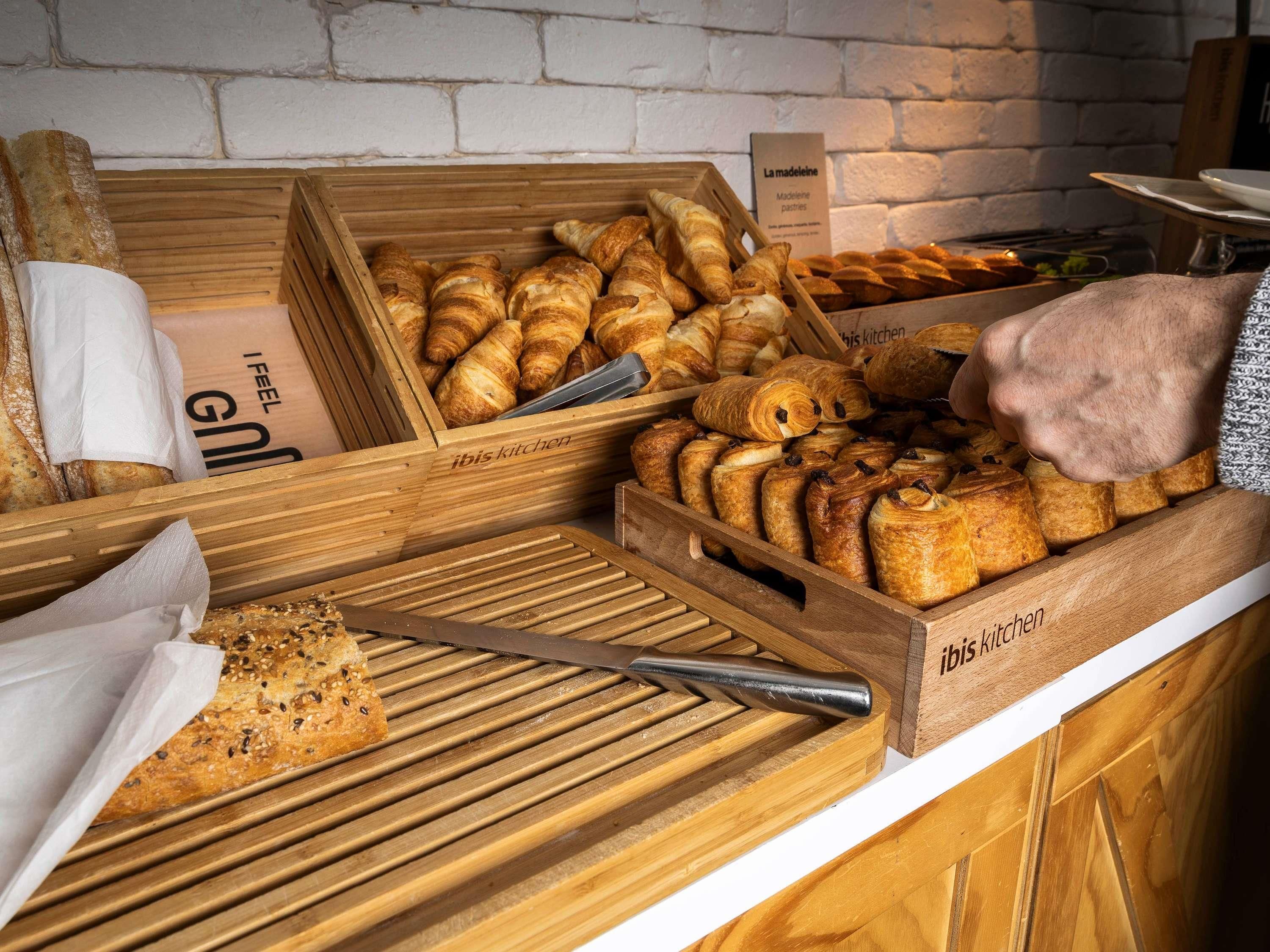Una vetrina di panetteria con diversi prodotti da forno, tra cui croissant e pane, in casse di legno etichettate "ibis kitchen".