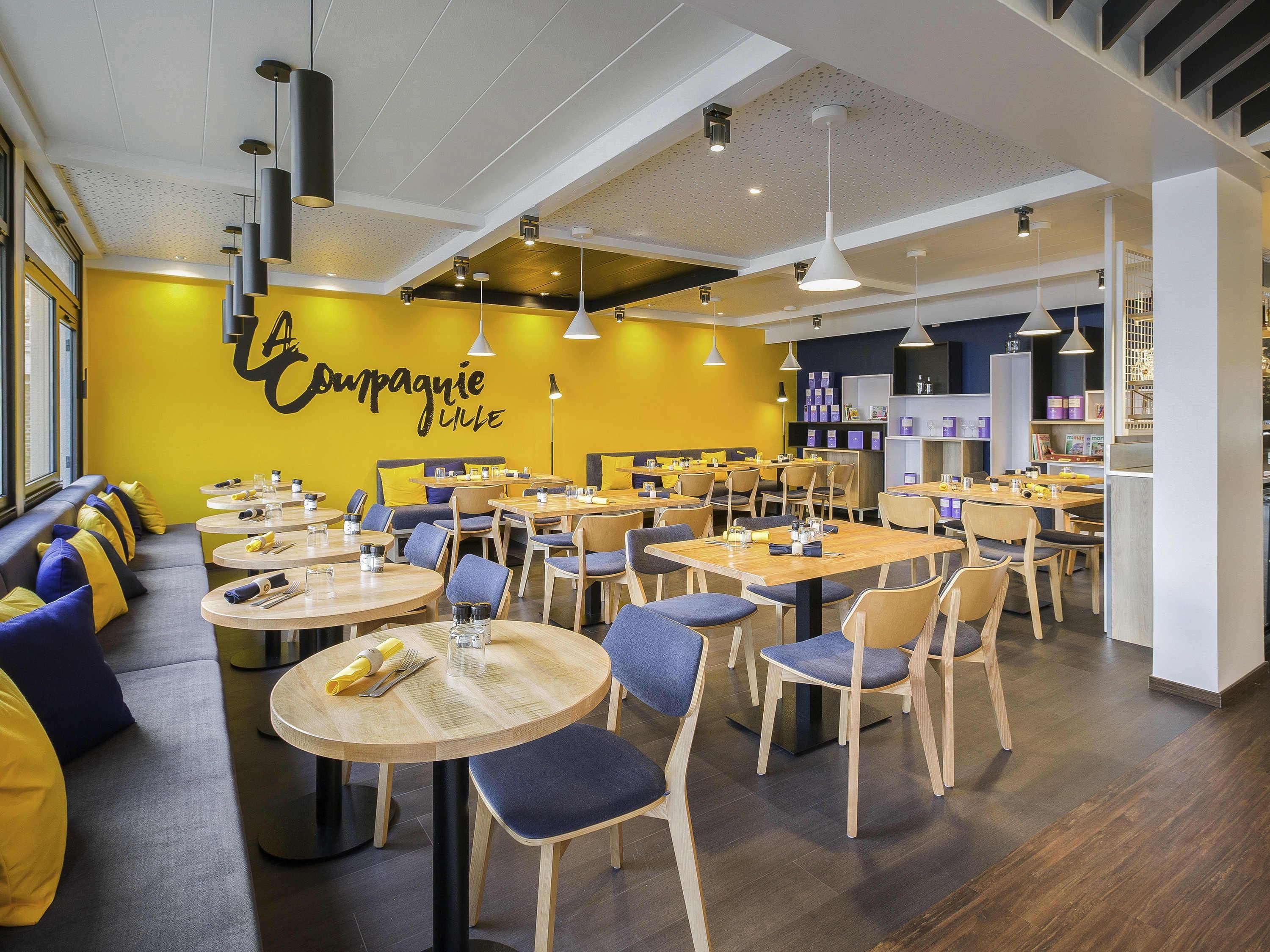 A modern restaurant interior with round tables, blue chairs, and a yellow accent wall.