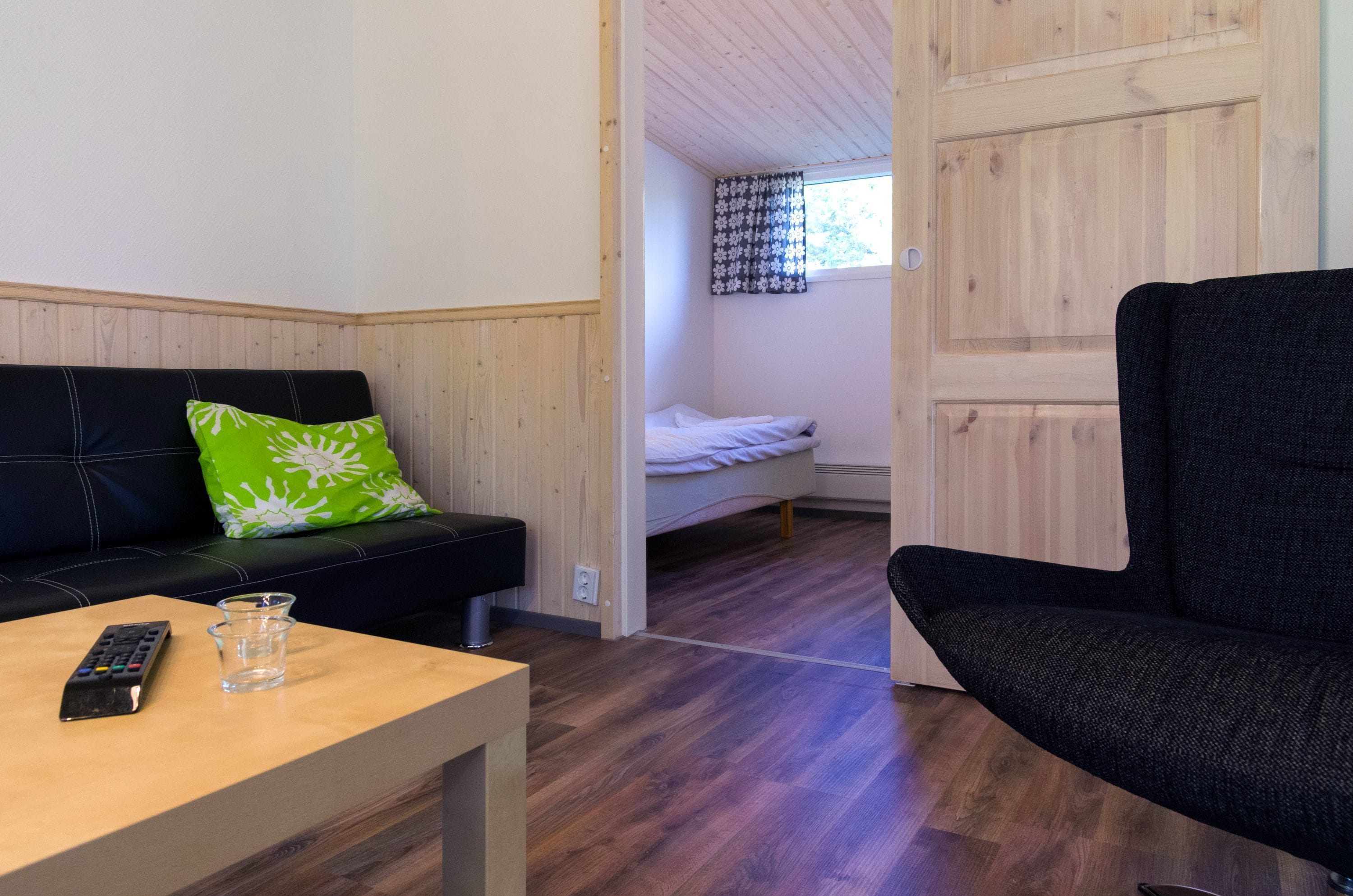 A room with a wooden floor, a black sofa with a green pillow, a bed with white bedding, and a black armchair.