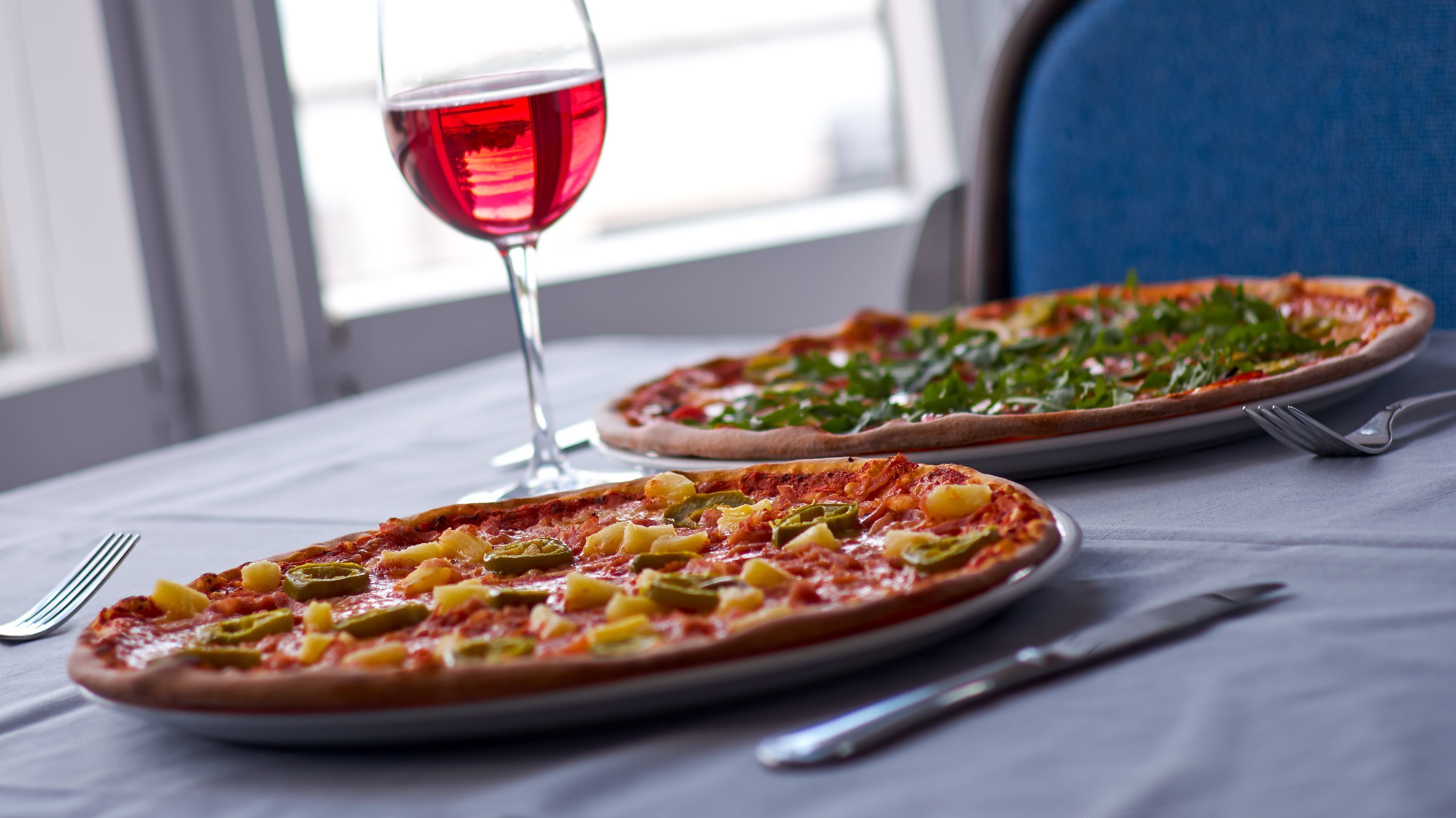 Two pizzas on plates with a glass of red wine on a table by a window.