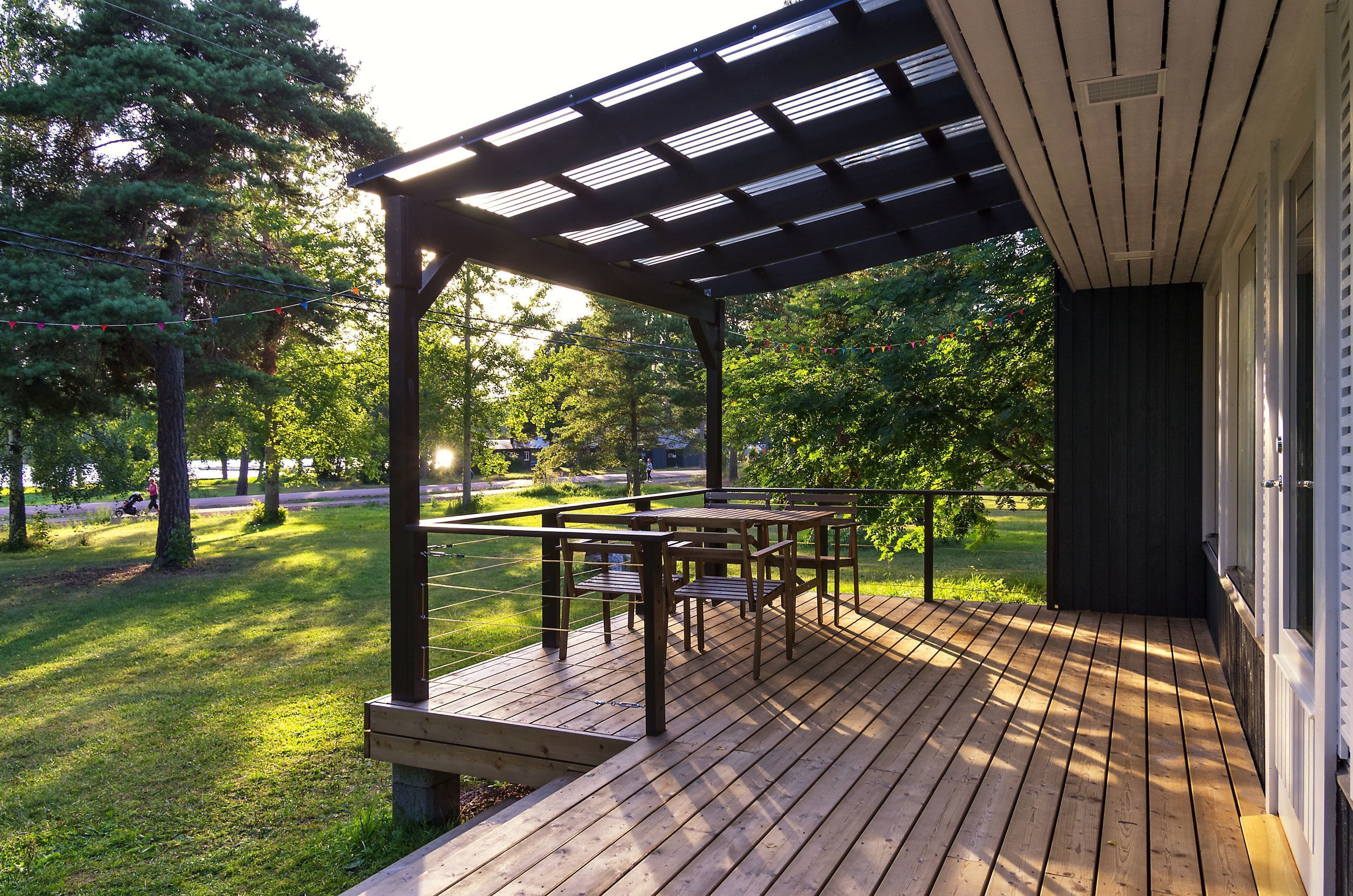 Una terraza de madera con un pérgola, mobiliario exterior y vistas a un parque con árboles y una carretera.