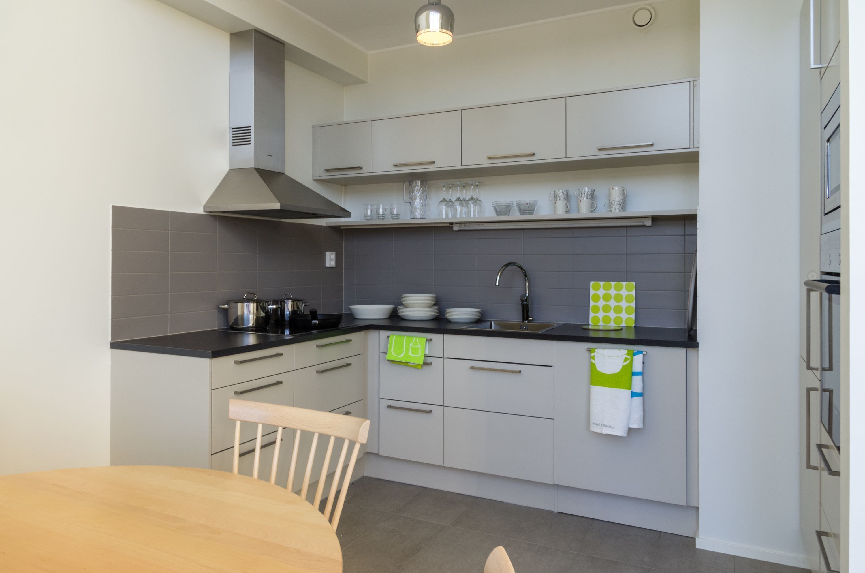 Eine moderne K üche mit grauen Schränken, einer schwarzen Arbeitsplatte und einer Edelstahl-Dunstabzugshaube. Ein Esstisch mit einem Stuhl steht bereit, und ein grün-weißes Handtuch hängt an den Schrankgriffen.