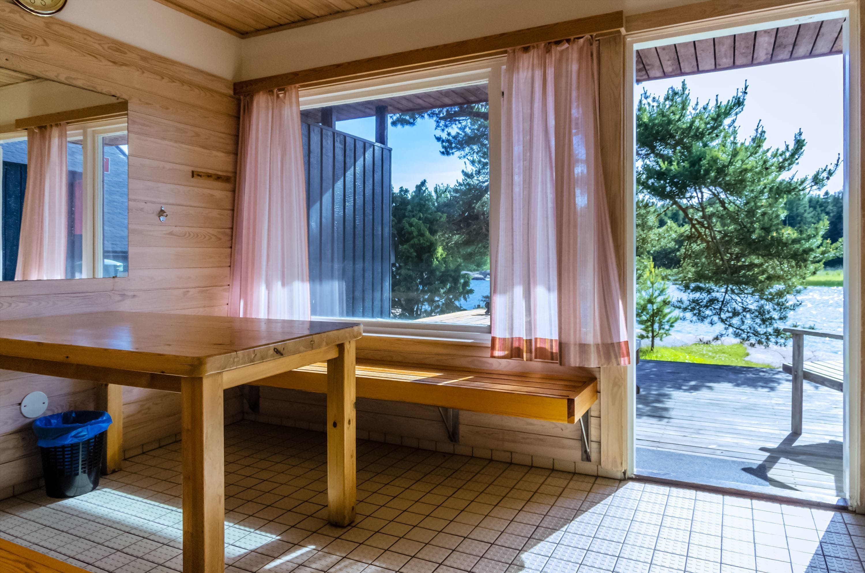 A wooden cabin with a large window, a wooden table, and a blue trash bin.