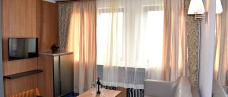 A hotel room with a wooden panel wall, a flat-screen TV, a minibar, a sofa, a coffee table, and a large window with sheer curtains.