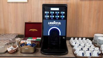 A coffee machine with multiple buttons and a display, a basket with coffee pods, a tray of cups, and a stack of plates.