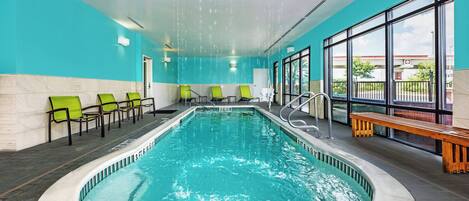 An indoor swimming pool with clear blue water, surrounded by green and black chairs, and a wooden bench.