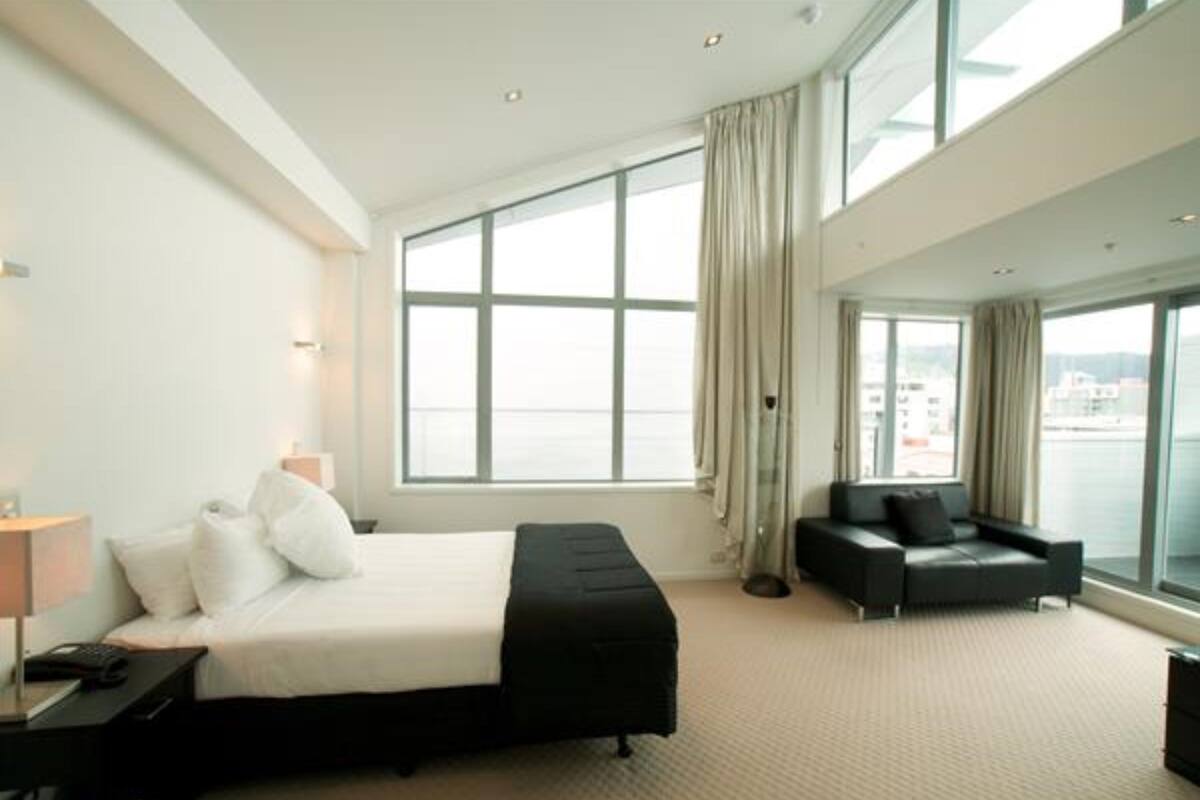 A modern hotel room with a large bed, a sofa, and a view of the city through large windows.