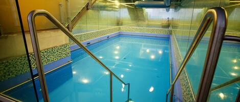 An indoor swimming pool with clear glass walls, metal railings, and tiled flooring.
