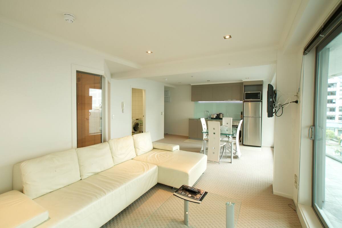 A modern living room with a sectional sofa, a dining area with a glass table, and a kitchen with stainless steel appliances.