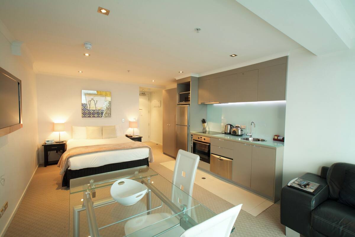A modern hotel room with a glass dining table, a bed with a white headboard, a kitchenette, and a black leather sofa.