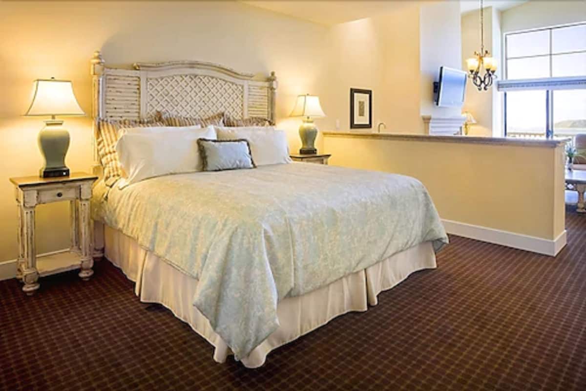 A neatly made bed with a headboard, two bedside tables with lamps, a large window, and a chandelier.