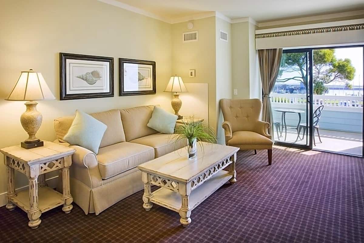 A hotel room with a beige sofa, two side tables with lamps, a coffee table, and a view of the outdoors through a large window.