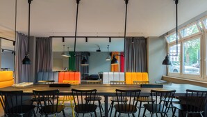A modern dining area with a long table and multiple chairs, a sofa with colorful cushions, and pendant lights. - a&o Düsseldorf Hauptbahnhof (Düsseldorf)