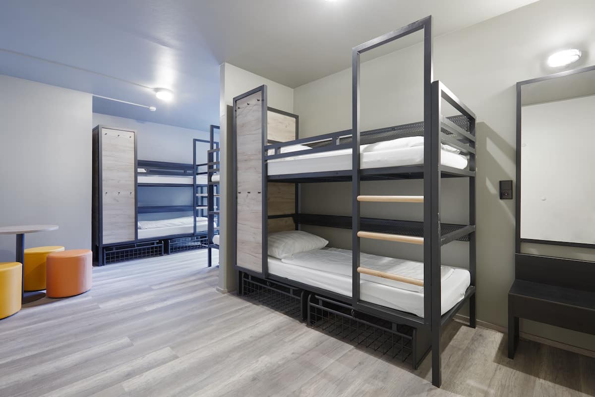 Dormitorio con literas, tres camas superpuestas, una mesita pequeña y dos taburetes naranjas.
