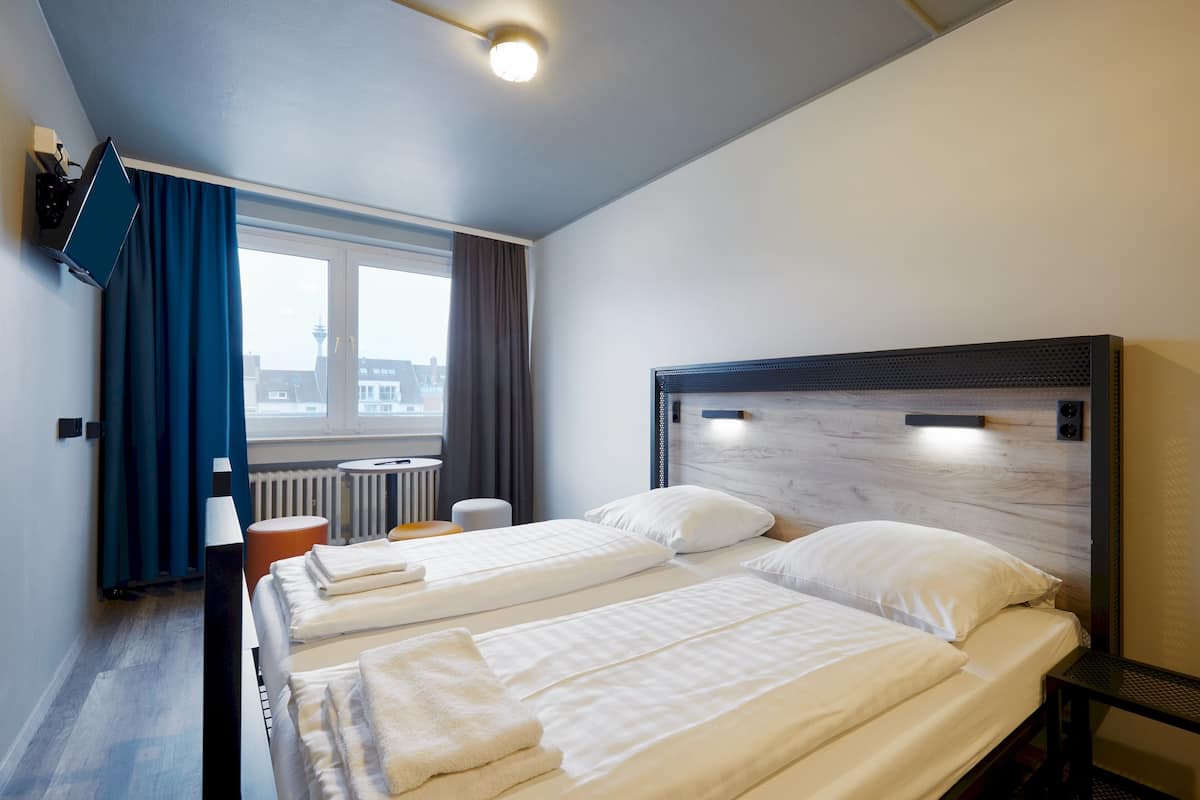 Habitación de hotel con dos camas, un televisor montado en la pared, una ventana con cortinas y vistas a edificios.