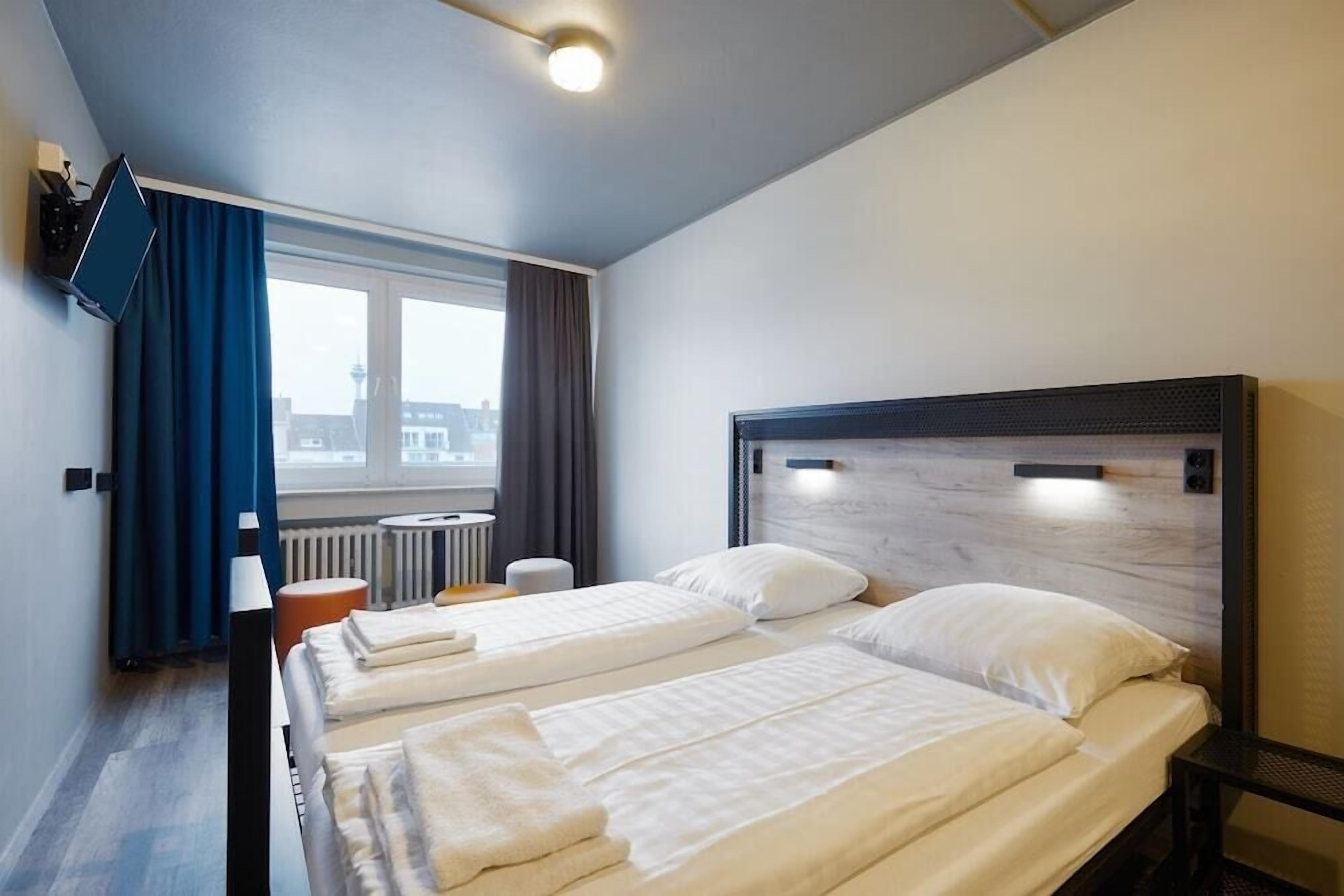 A hotel room with two beds, a TV mounted on the wall, a window with curtains, and a radiator.