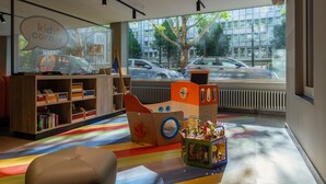 A children's play area with colorful play structures and a large window offering a view of parked cars and trees. - a&o Düsseldorf Hauptbahnhof (Düsseldorf)