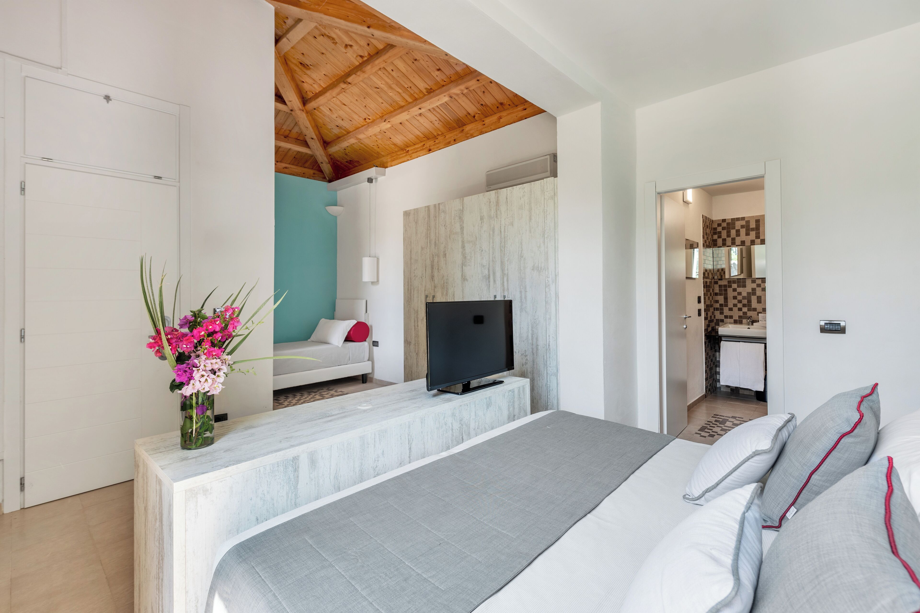 Un dormitorio con cama, televisor, escritorio pequeño con un florero y baño visible a través de una puerta abierta.