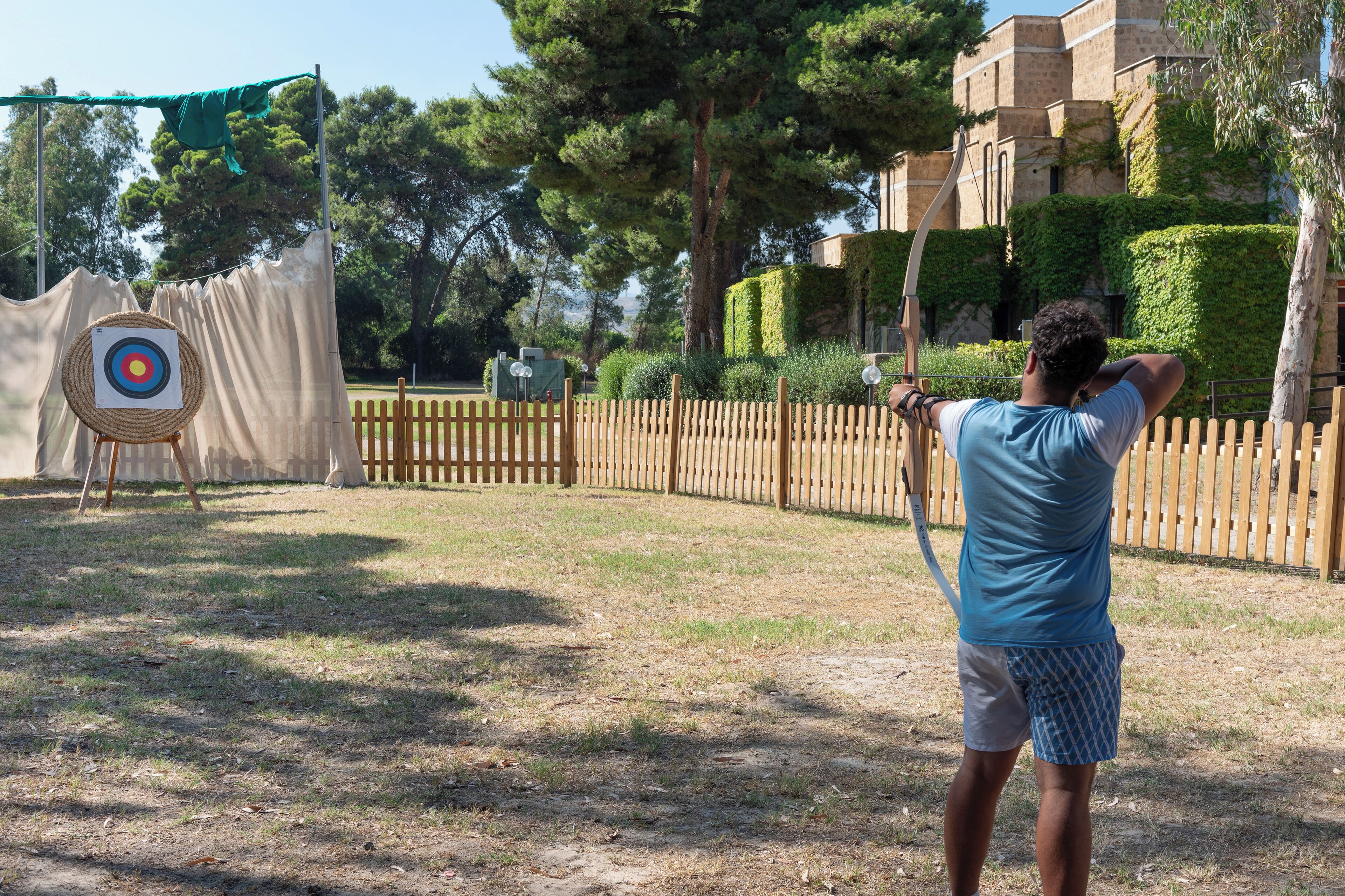 Un homme vise une cible d’archerie dans un cadre extérieur, avec une clôture en bois et de la verdure en arrière-plan.