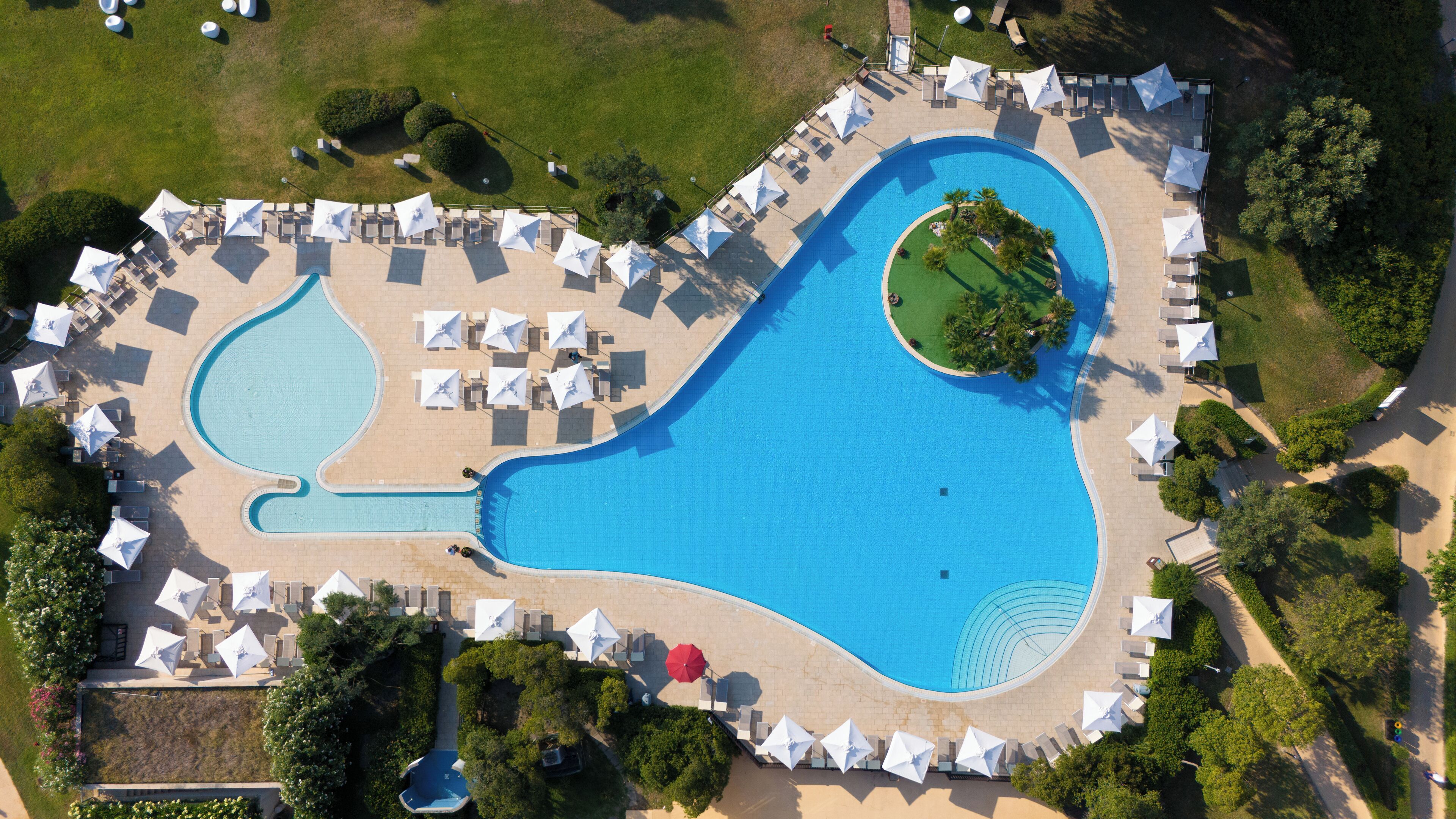 An aerial view of a resort with a large swimming pool, sun loungers, and umbrellas.