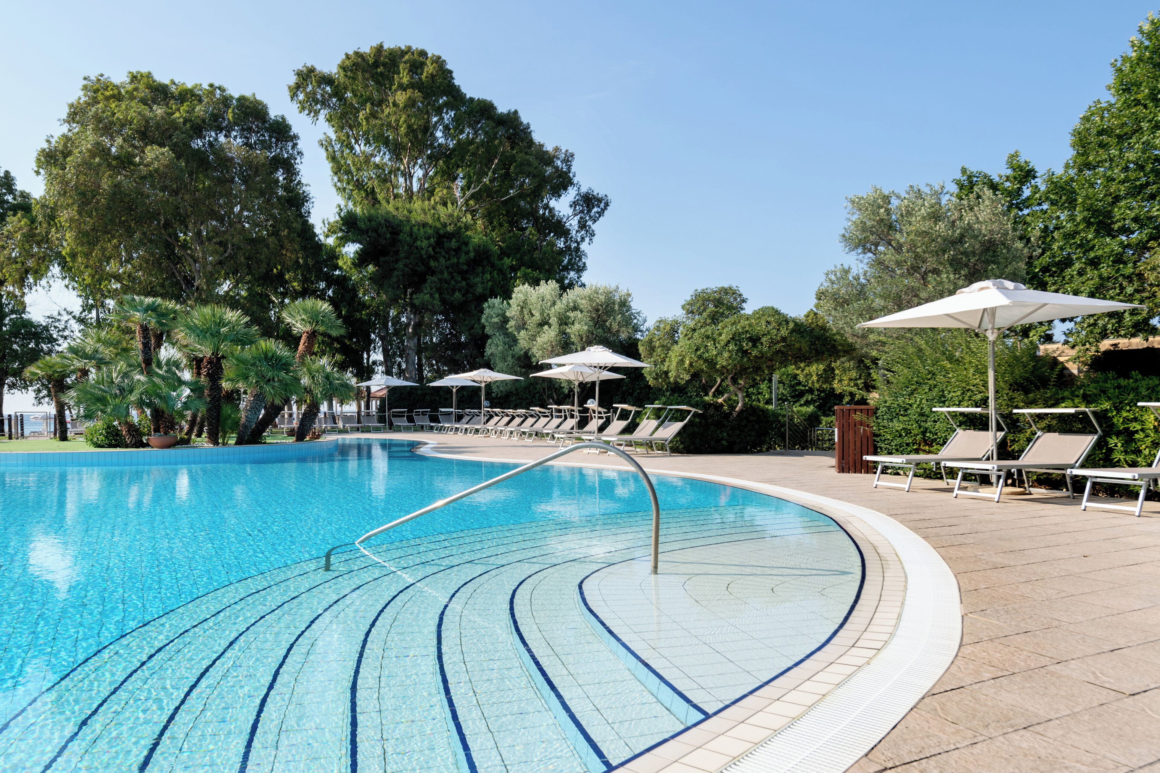 A swimming pool with clear blue water, surrounded by lounge chairs and umbrellas, and trees in the background.