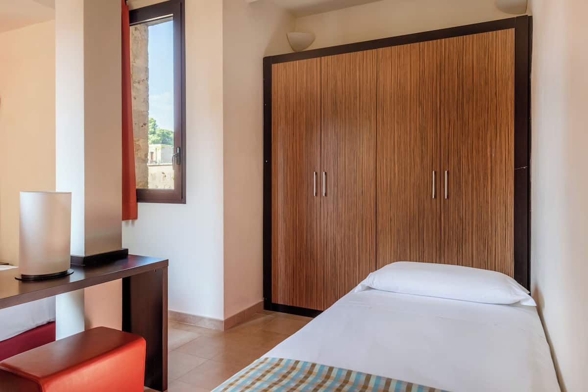 A hotel room with a bed, a desk with a lamp, a large wardrobe, and a window with a view of the outside.