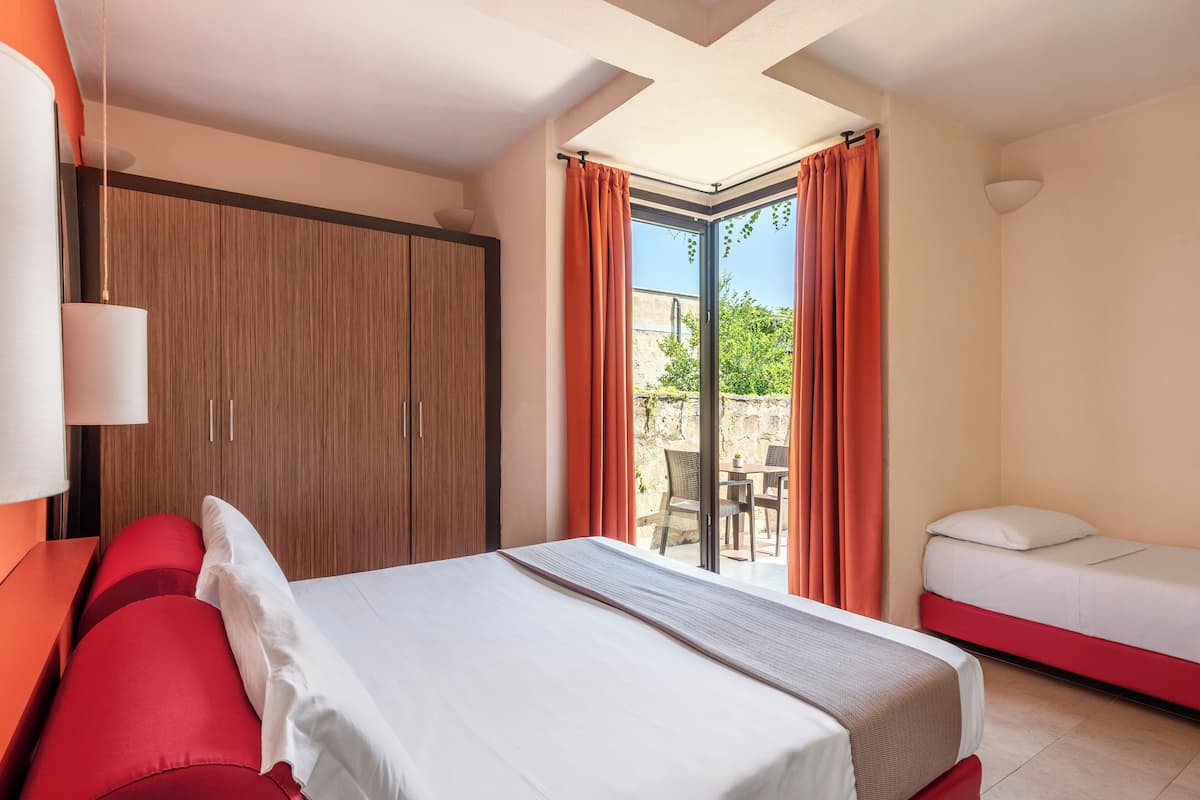 A hotel room with a bed, a red sofa, a wooden wardrobe, and a sliding glass door to the outside.