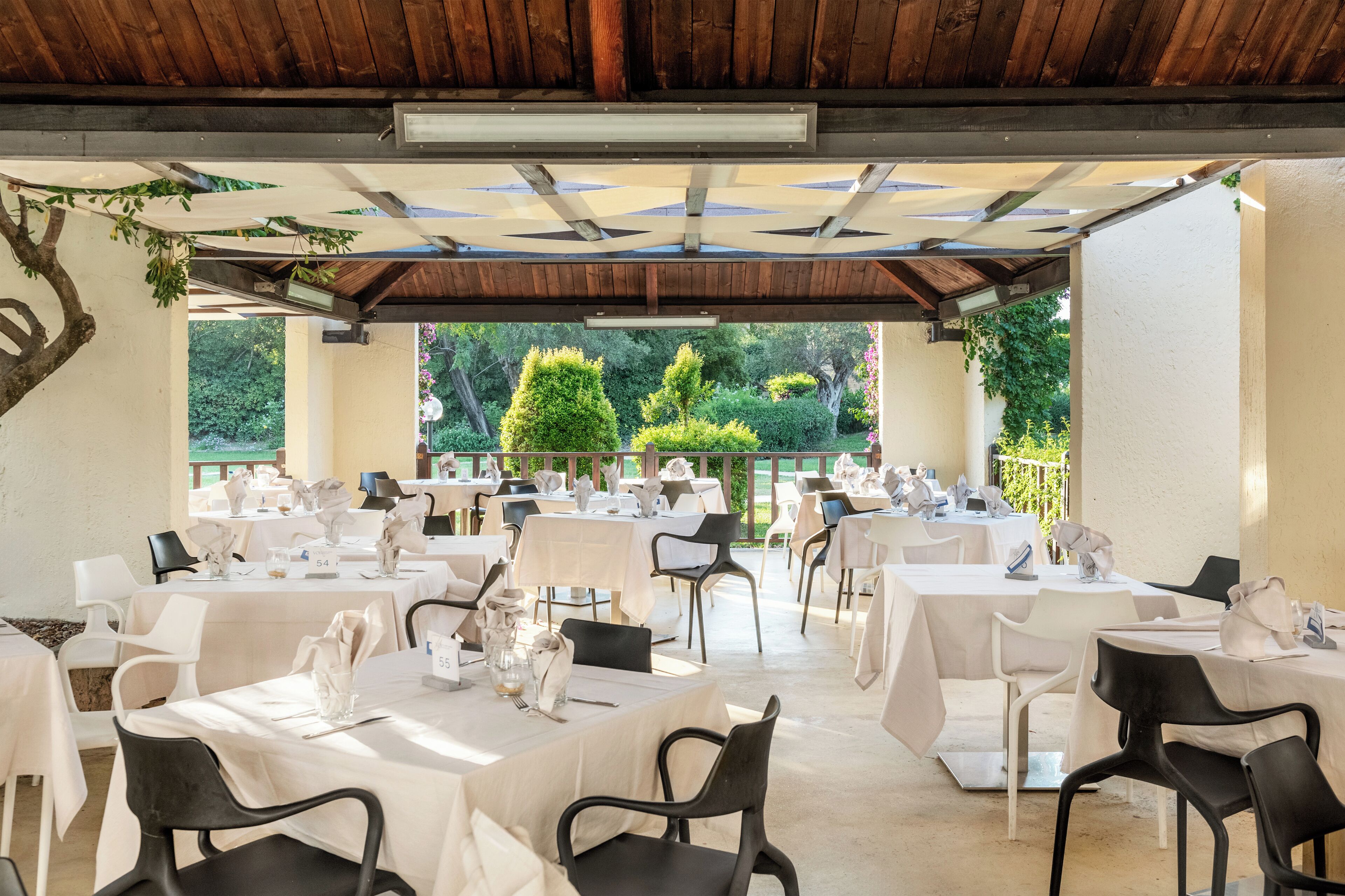 A covered outdoor dining area with tables set for a meal, surrounded by greenery and a wooden ceiling.