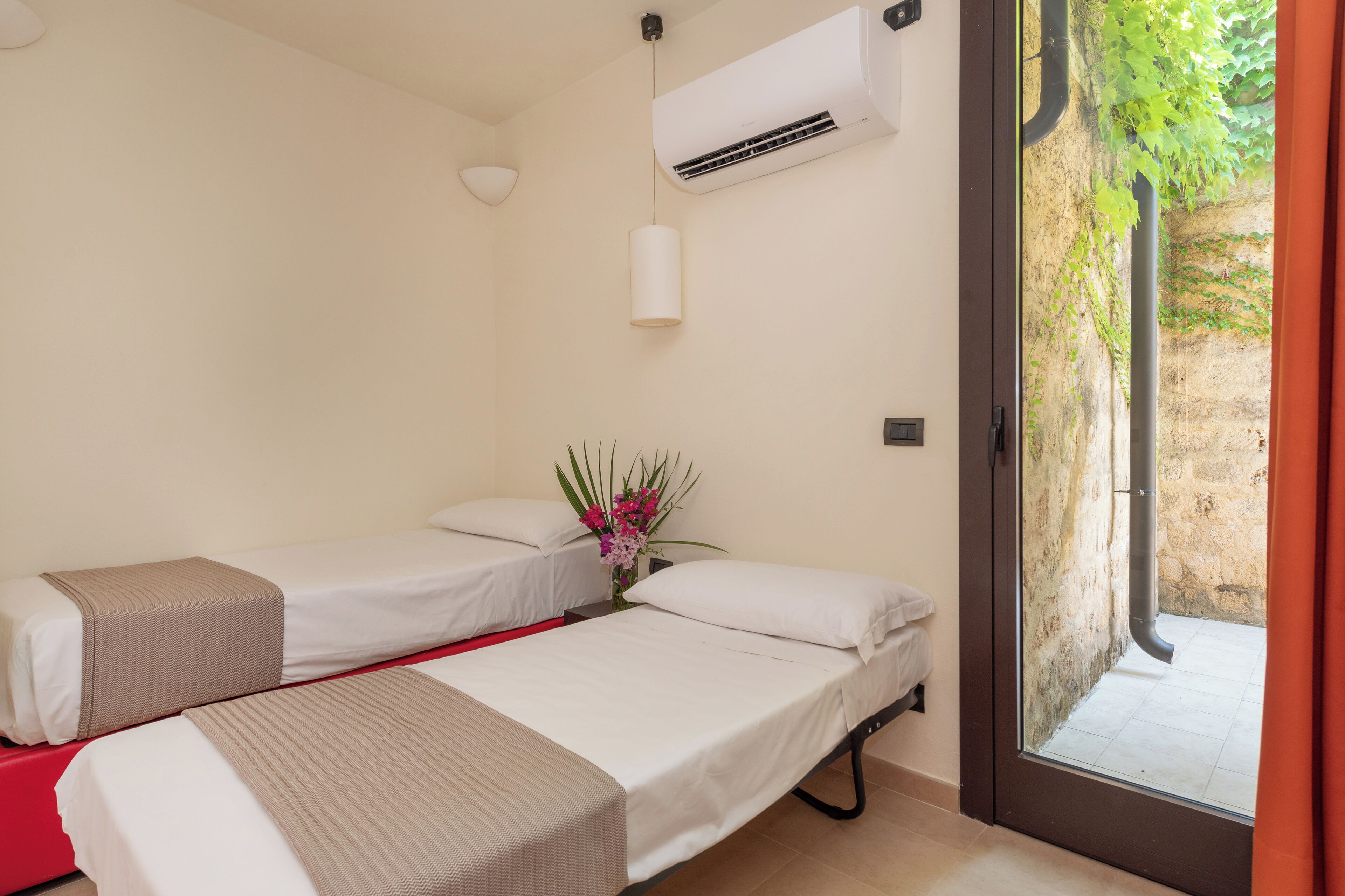 A hotel room with two single beds, a small table with a vase of flowers, an air conditioning unit, and a view of a stone wall and greenery outside.