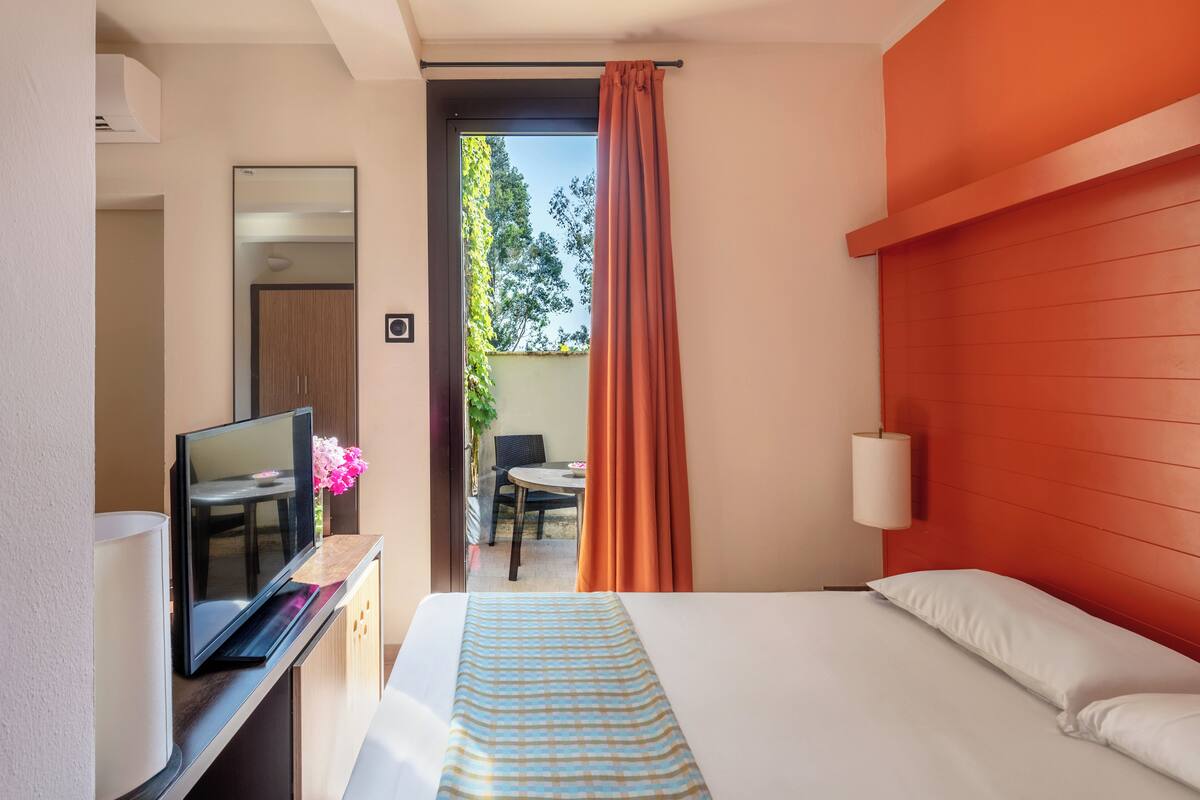 A hotel room with a bed, a TV on a desk, a small table with chairs, and a view of the outdoors.