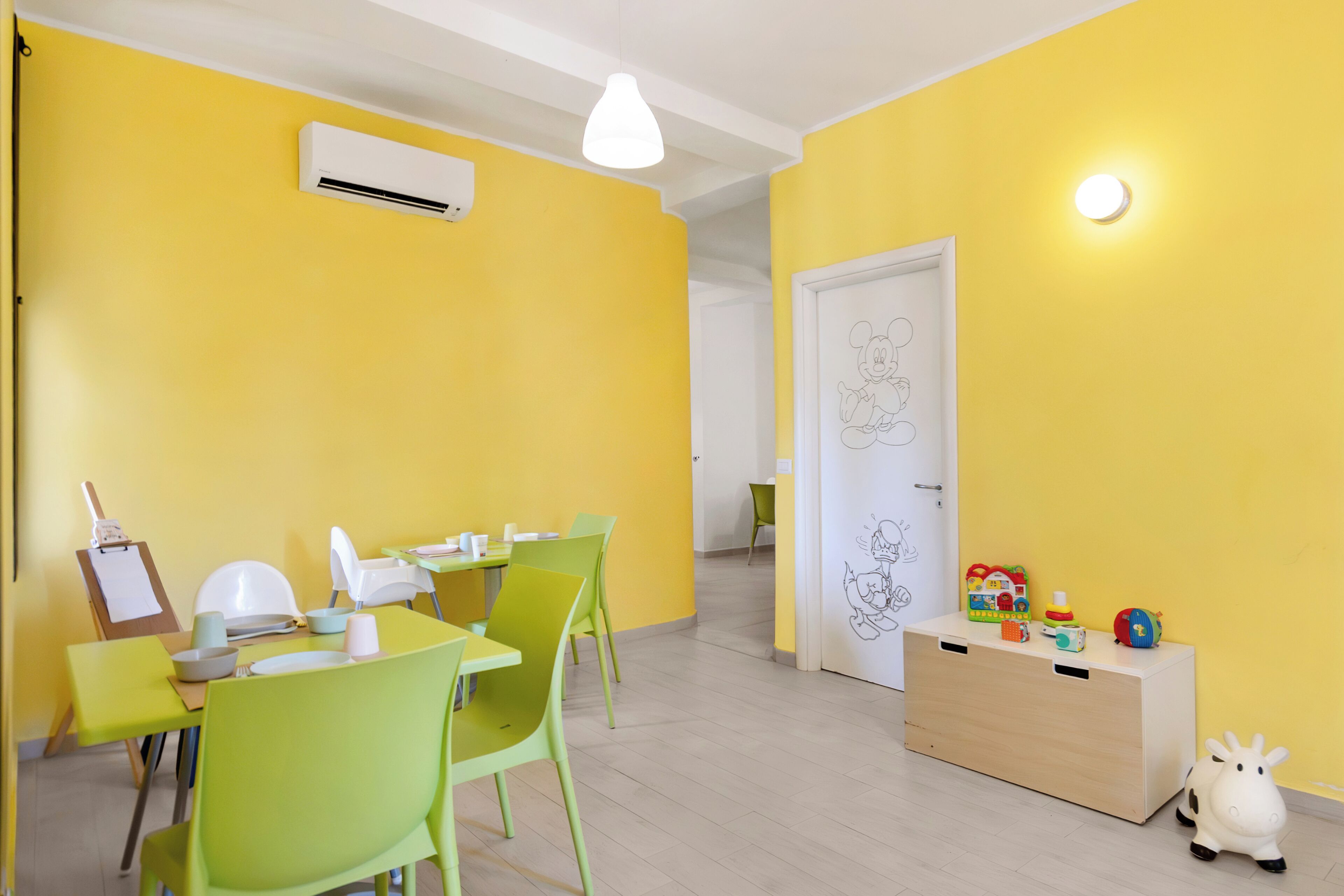 Une pièce lumineuse aux murs jaune vif, avec une table à manger, des chaises et un espace de jeux équipé de jouets.