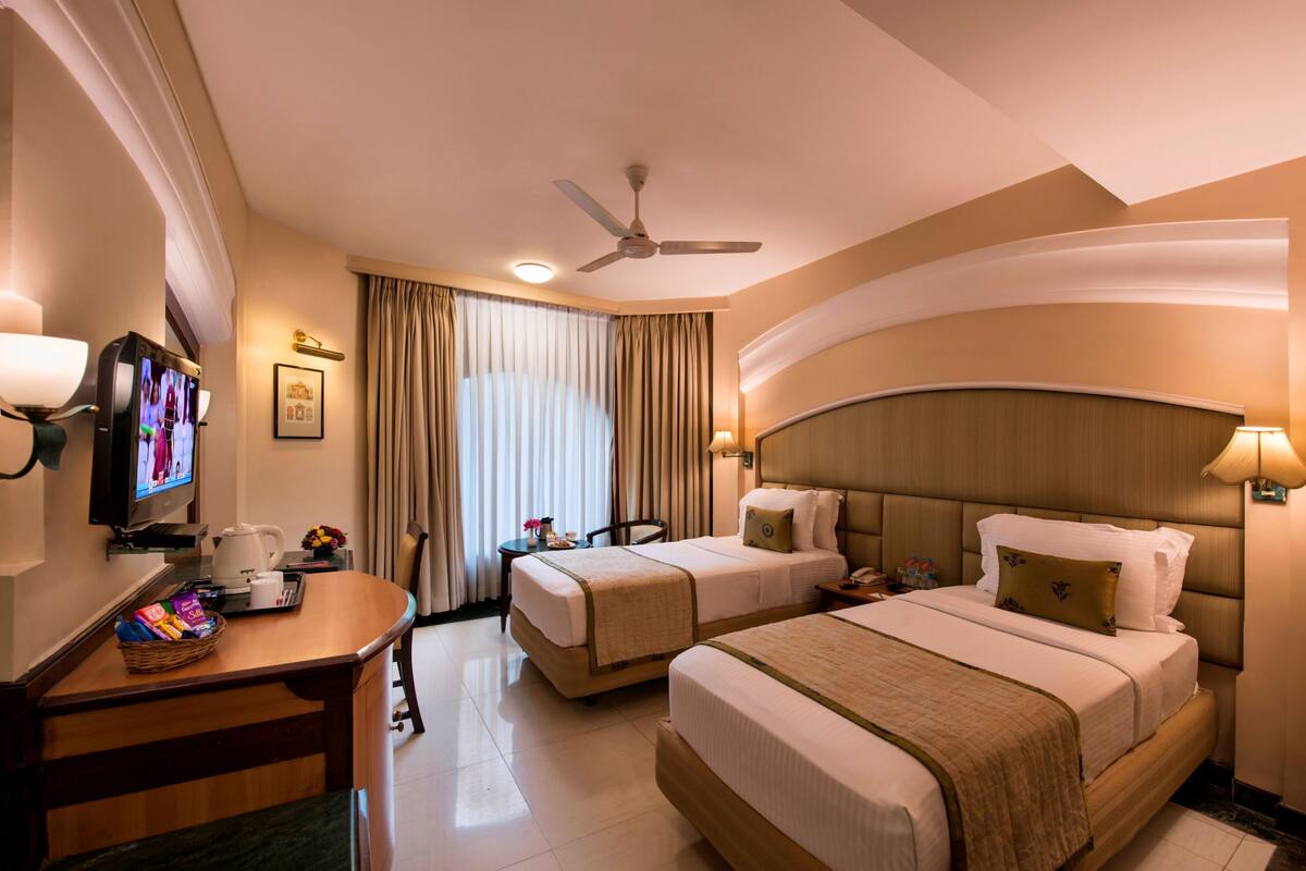 A hotel room with two beds, a desk with a television, a ceiling fan, and a large window with curtains.