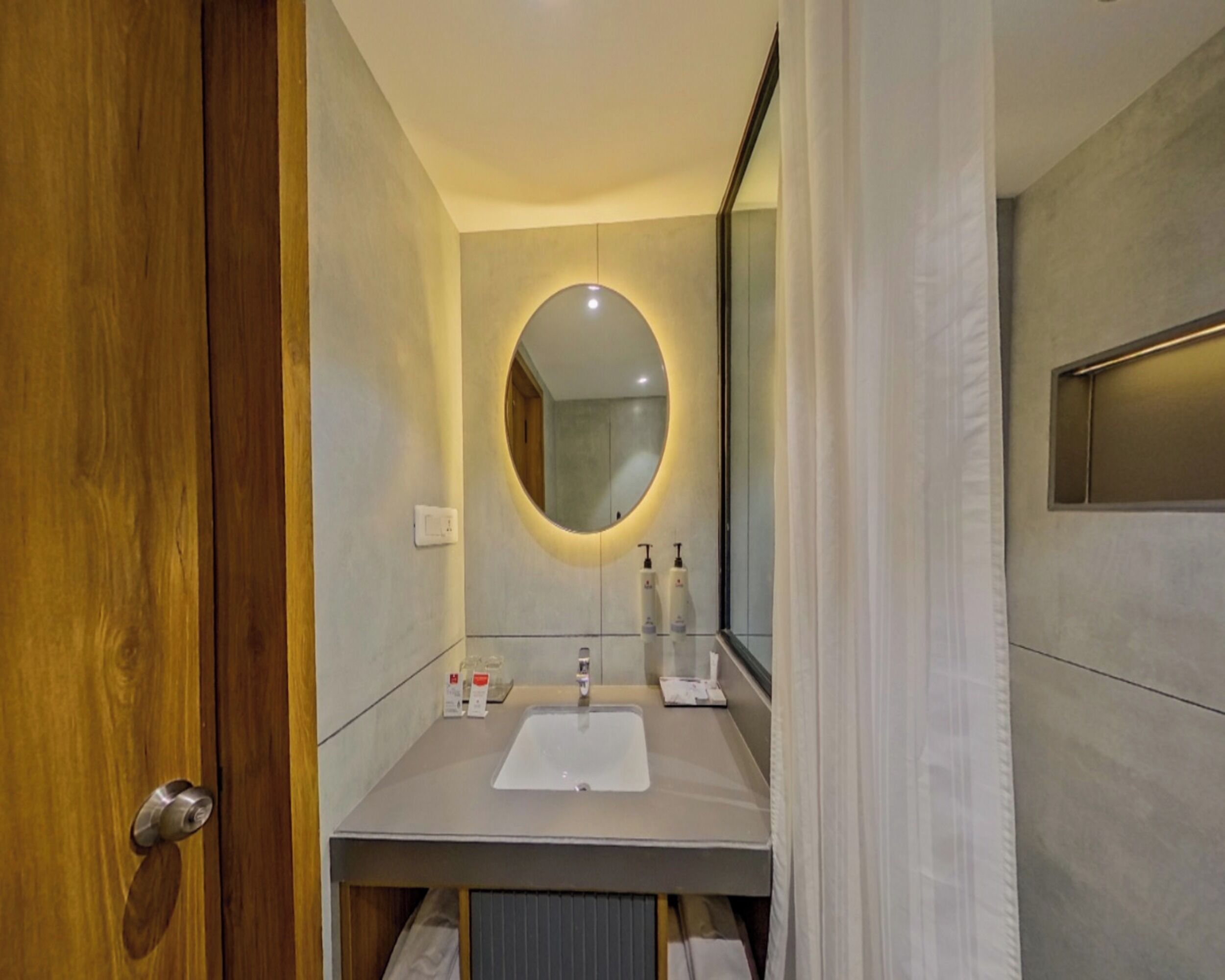 Un baño moderno con puerta de madera, espejo redondo, lavamanos blanco y artículos de aseo.
