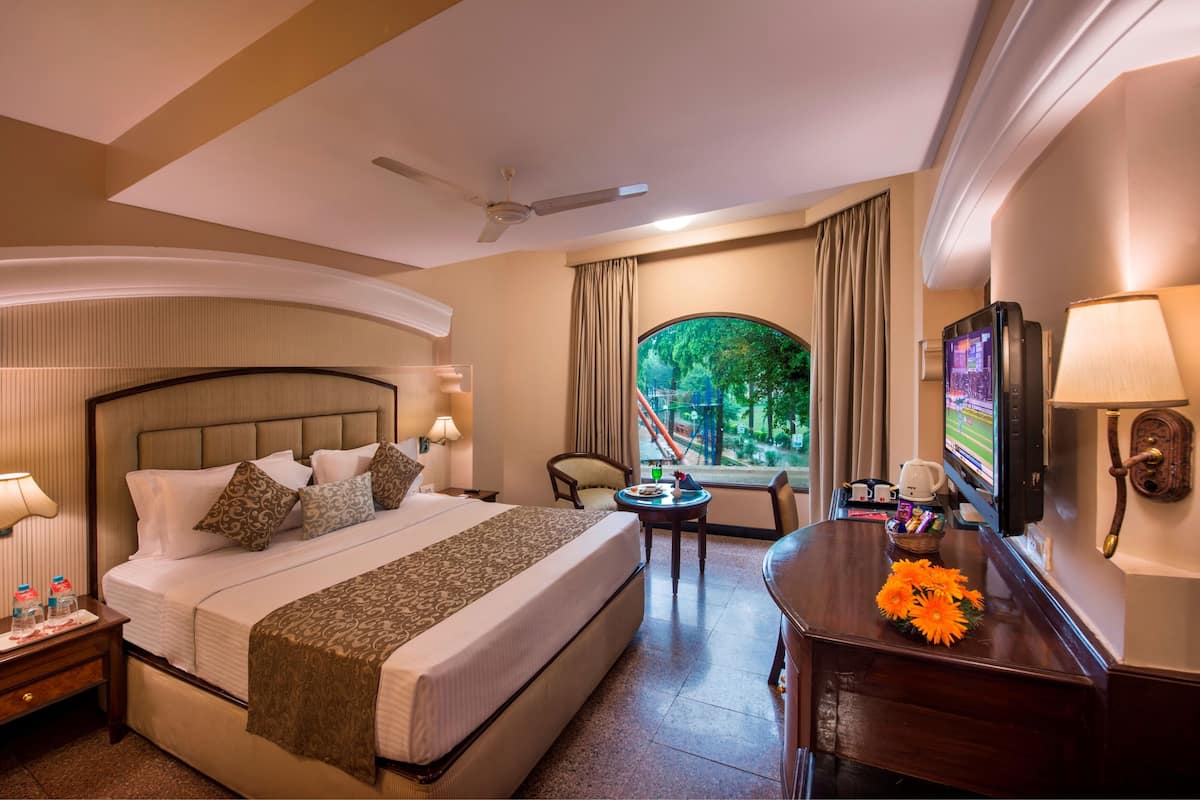 A hotel room with a bed, a desk, a chair, a television, and a window with a view of greenery.