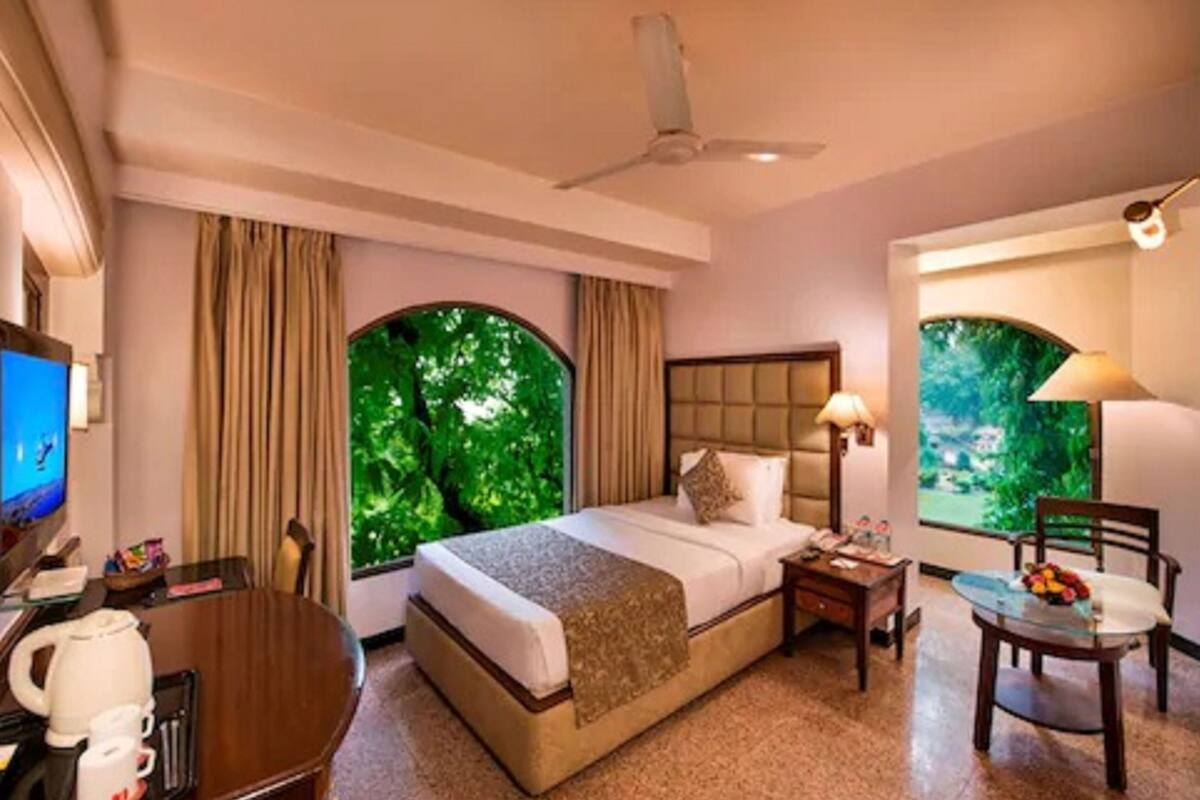 A hotel room with a large bed, a desk with a chair, a TV, and two windows with a view of greenery.