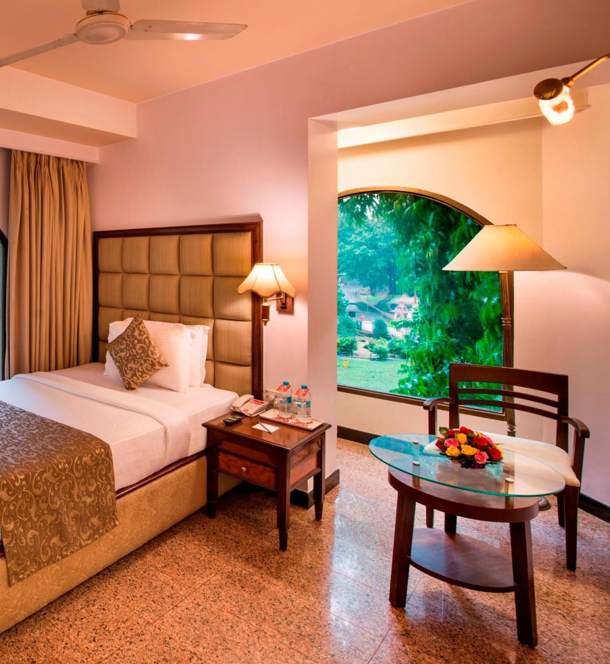 A hotel room with a bed, bedside table, lamp, chair, and a view of greenery through a window.