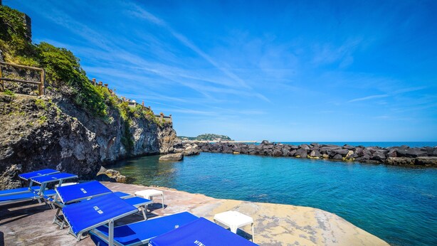 A beachside hotel with blue sun loungers and a rocky cliff. - Albergo Terme Italia (Casamicciola Terme)