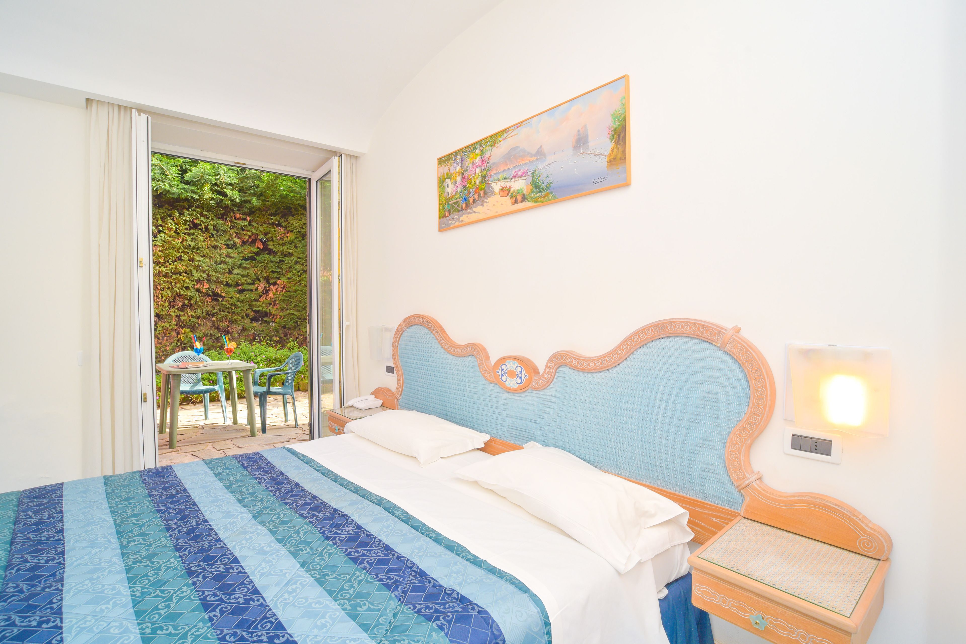 Ein Schlafzimmer mit Bett, Nachttisch, einem Gemälde und Blick auf eine Terrasse mit Tisch und Stühlen.