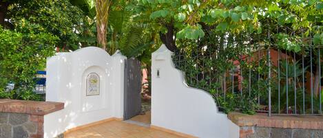 A gated entrance with a white wall and a sign, surrounded by lush greenery and a stone wall.